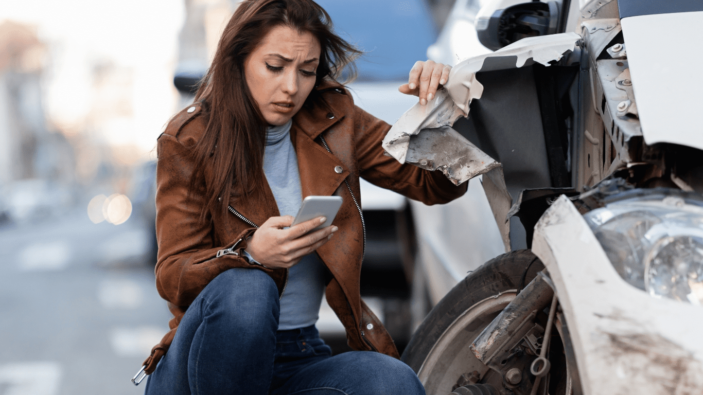 A woman who wants to save money on autobody repair kneels next to her damaged car, looking at her phone.